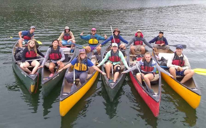 a group of students in six canoes smile for a photo on calm water